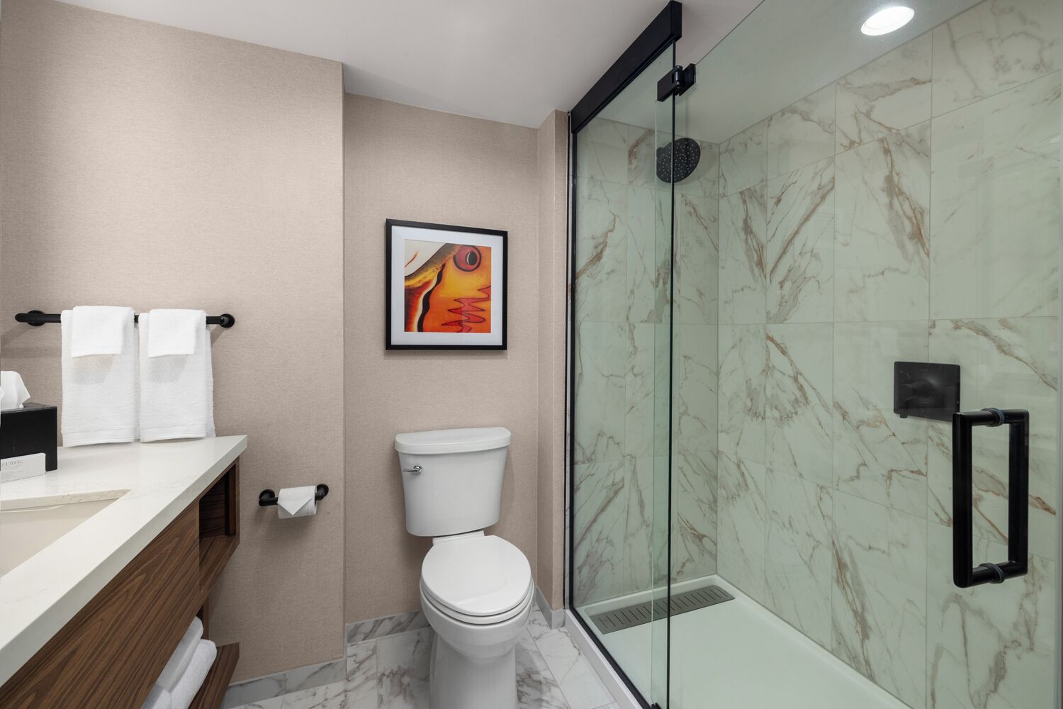 Un baño moderno con ducha de vidrio, inodoro, lavamanos con toallero y una obra de arte enmarcada en la pared.
