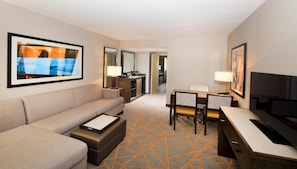 Habitación de hotel con sofá, escritorio, televisión, mesa de comedor con sillas, zona de cocina y obras de arte en las paredes.