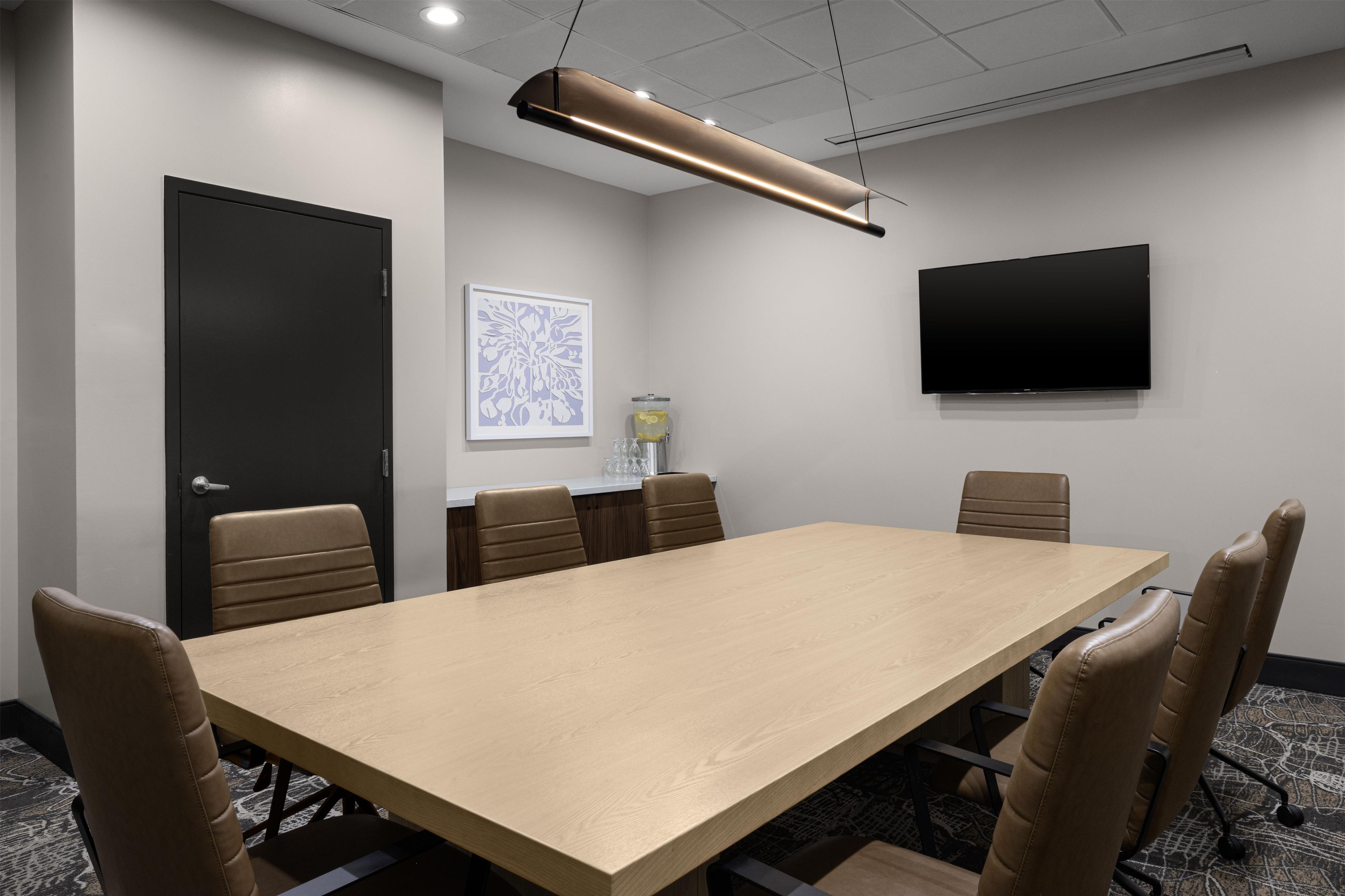 A conference room with a long table, six chairs, a flat-screen TV, and a framed artwork on the wall.