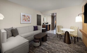 A modern hotel room with a sofa, a small table, a chair, a kitchen area, and a bathroom visible through an open door.