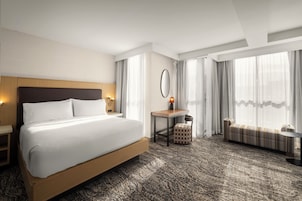 Habitación de hotel con una cama grande, un escritorio con silla, una mesita de noche con lámpara, un sofá y un ventanal con cortinas.