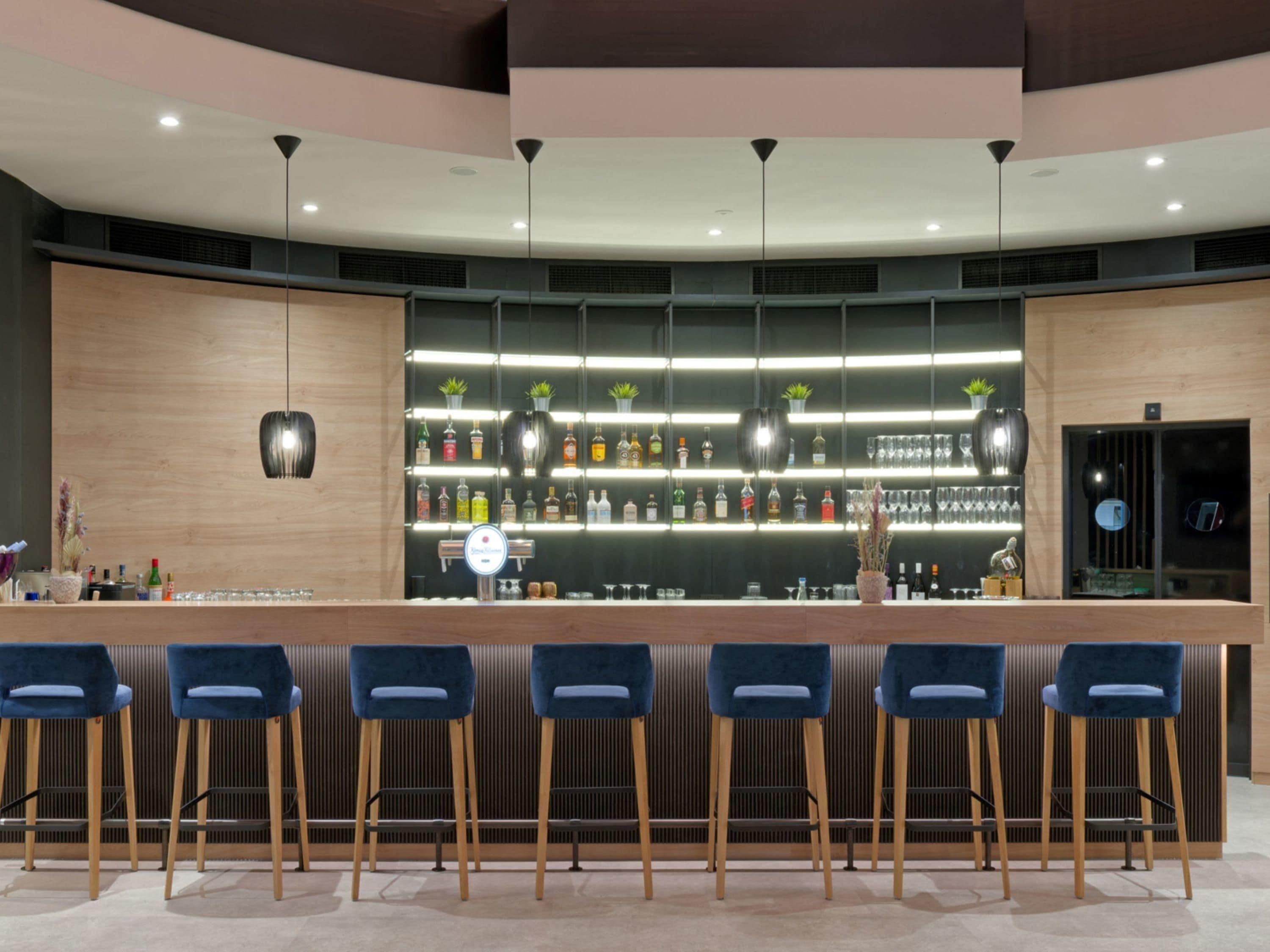 Zona de bar con mostrador, taburetes, estanterías con botellas y luces colgantes.