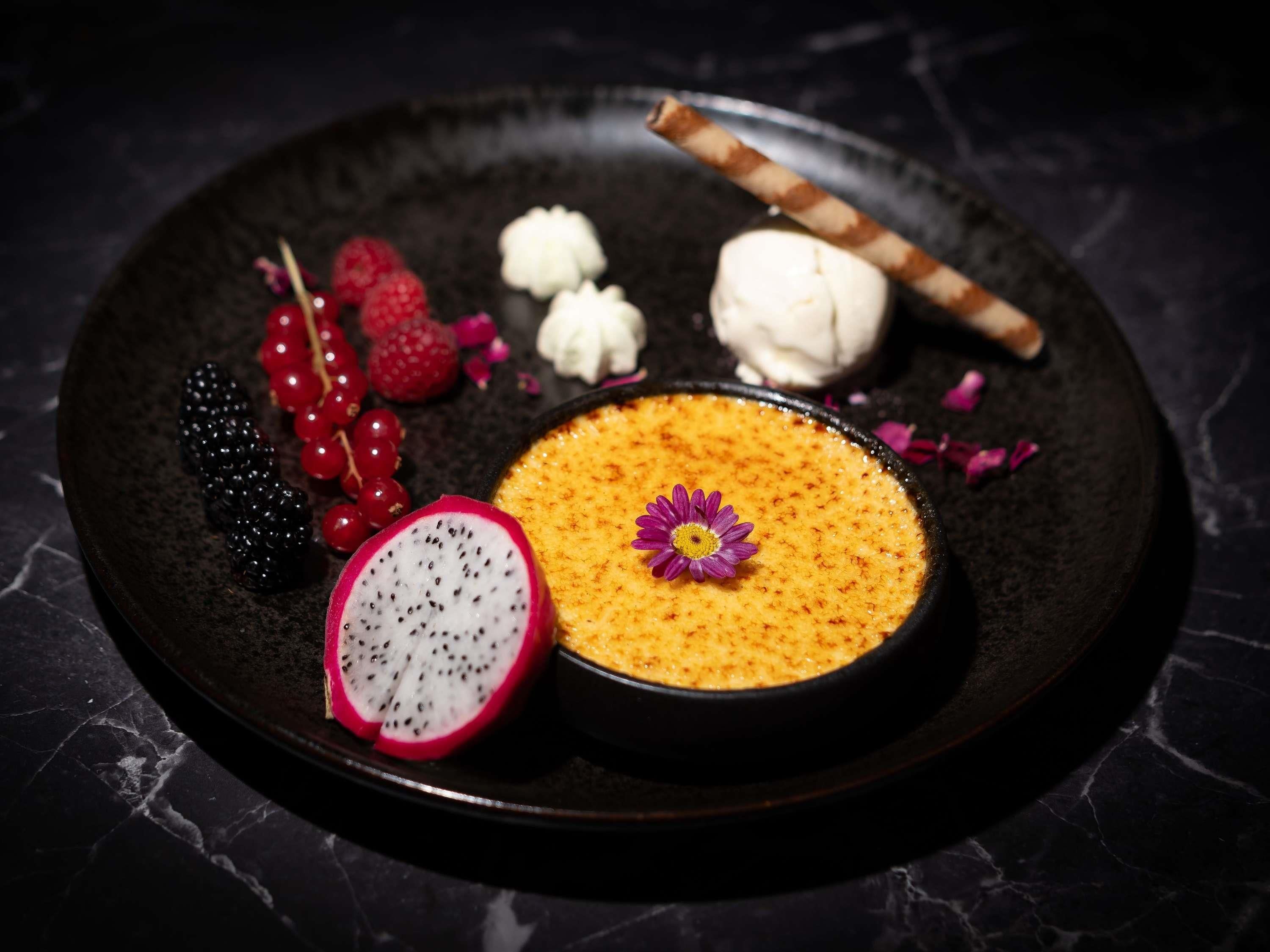 Un plat de postres amb un postre cremós, una làmina de pitaya, nabes, groselles i una flor.