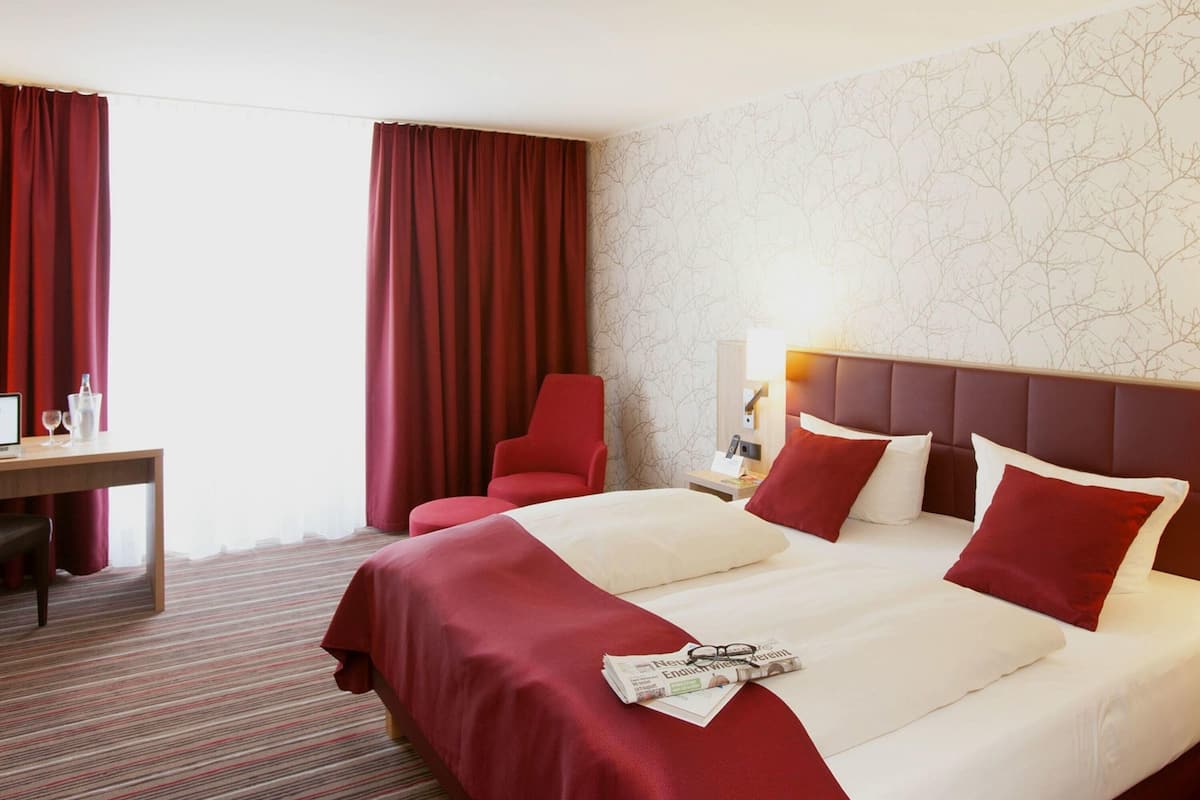 A hotel room with a large bed, two red chairs, a wooden desk, and a nightstand with a lamp.