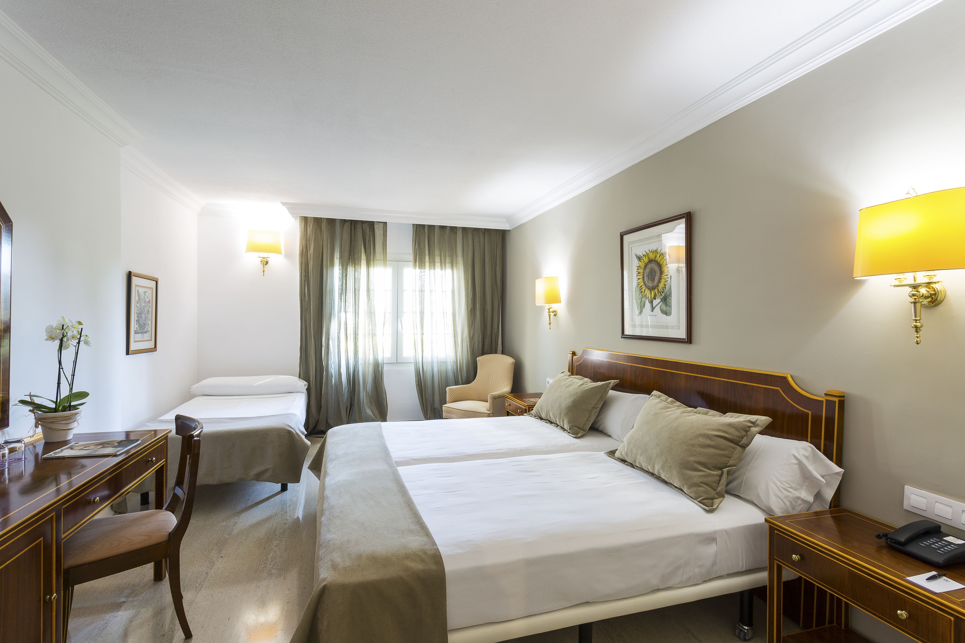 A hotel room with two beds, a desk, a chair, a bedside table, and a lamp.