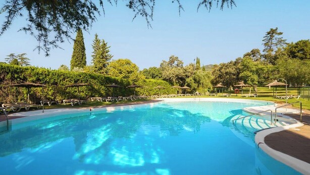 Una piscina cristalina rodeada de sillones de playa y sombrillas, con árboles y un cielo despejado al fondo.