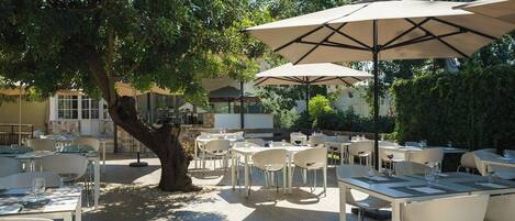 An outdoor dining area with tables and chairs, shaded by large umbrellas, and surrounded by trees.