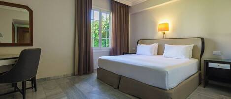 A hotel room with a bed, a desk, a chair, a nightstand, a lamp, and a window with curtains.