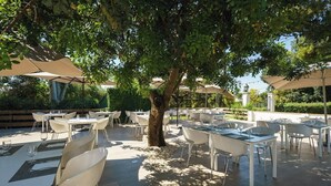 Un área de comedor al aire libre con mesas y sillas bajo sombrillas grandes, rodeada de árboles.