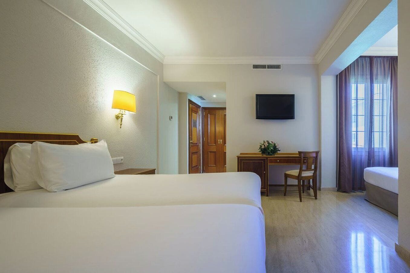 Ein Hotelzimmer mit einem großen Bett, einem Schreibtisch, einem Sessel, einem Fernseher und einem Fenster mit Vorhängen.