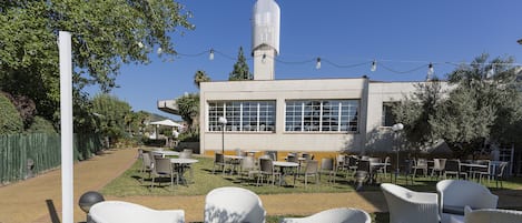 Un espacio al aire libre con sillas y mesas de mimbre, un edificio con grandes ventanales y una construcción alta con una cruz en la parte superior.