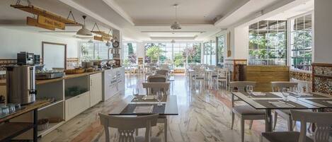 A spacious dining area with marble flooring, white furniture, and large windows offering a view of greenery outside.