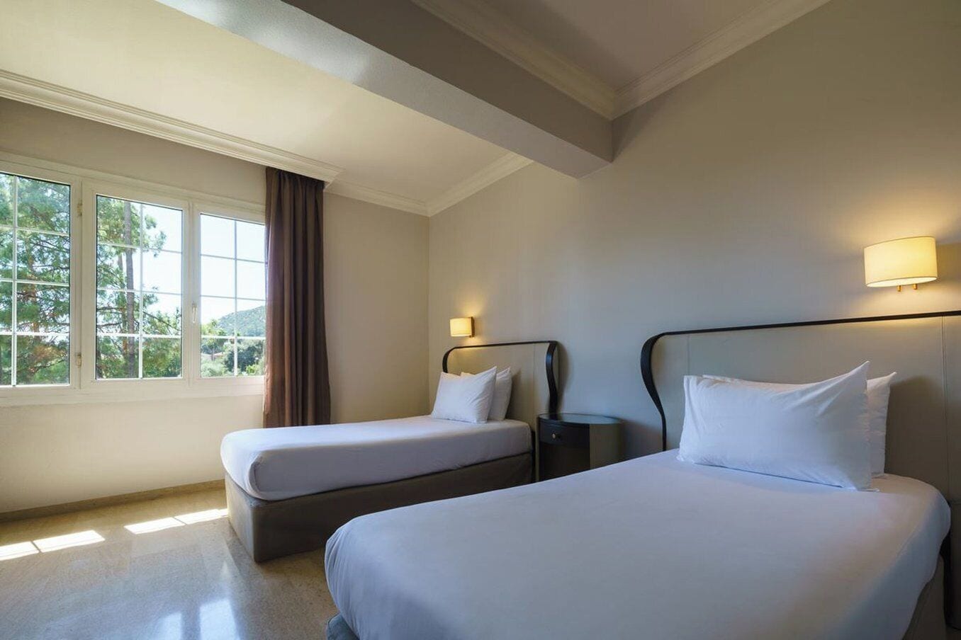 A hotel room with two beds, a window with curtains, and a nightstand with a lamp.