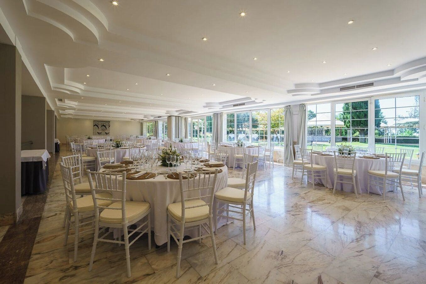 Un amplio salón de banquetes con mesas redondas y sillas blancas, grandes ventanales con vistas al exterior y suelo de mármol.
