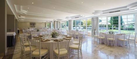 Un amplio salón de banquetes con mesas redondas y sillas blancas, grandes ventanales con vistas al exterior y suelo de mármol.
