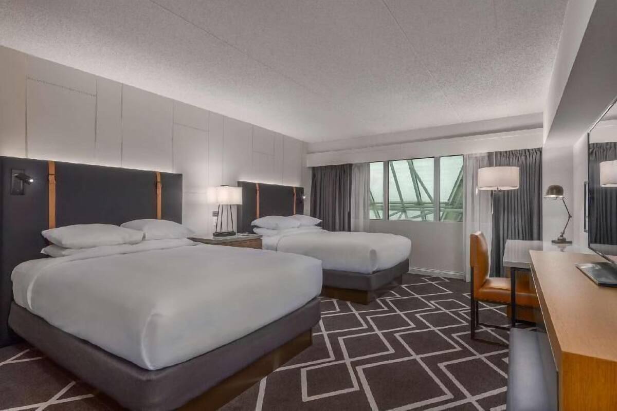 A hotel room with two beds, a desk, a chair, a lamp, and a view of the outside.