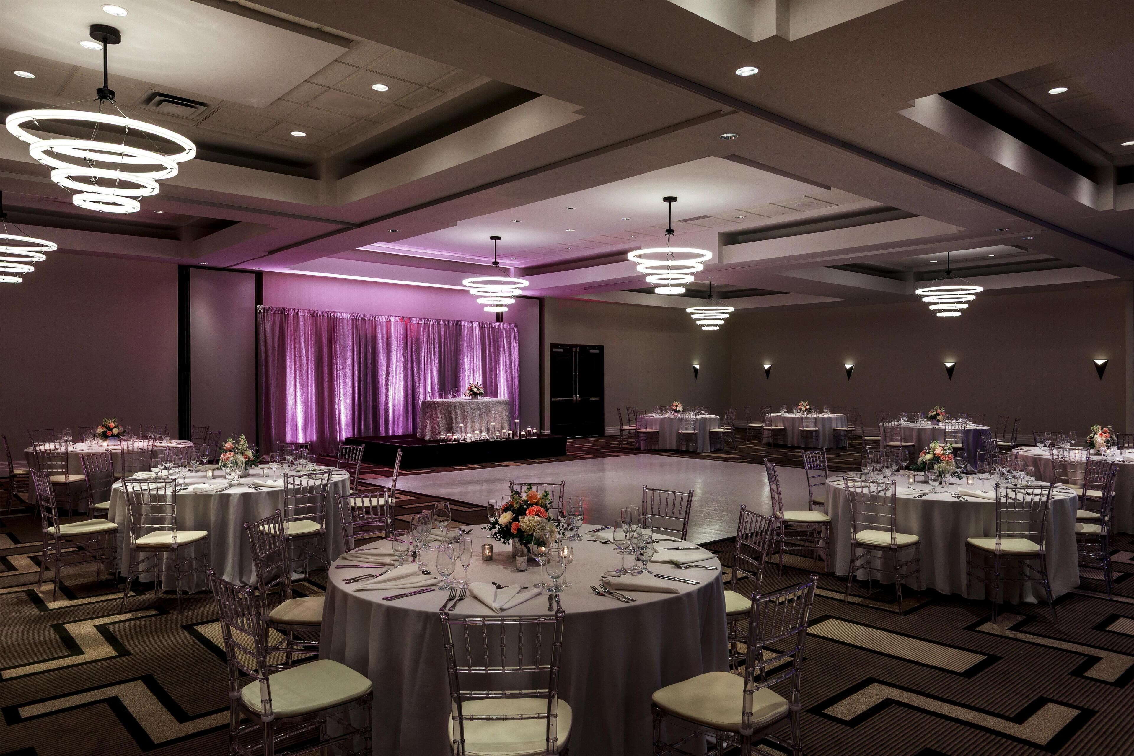 A banquet hall with multiple round tables, clear chairs, and modern pendant lighting.