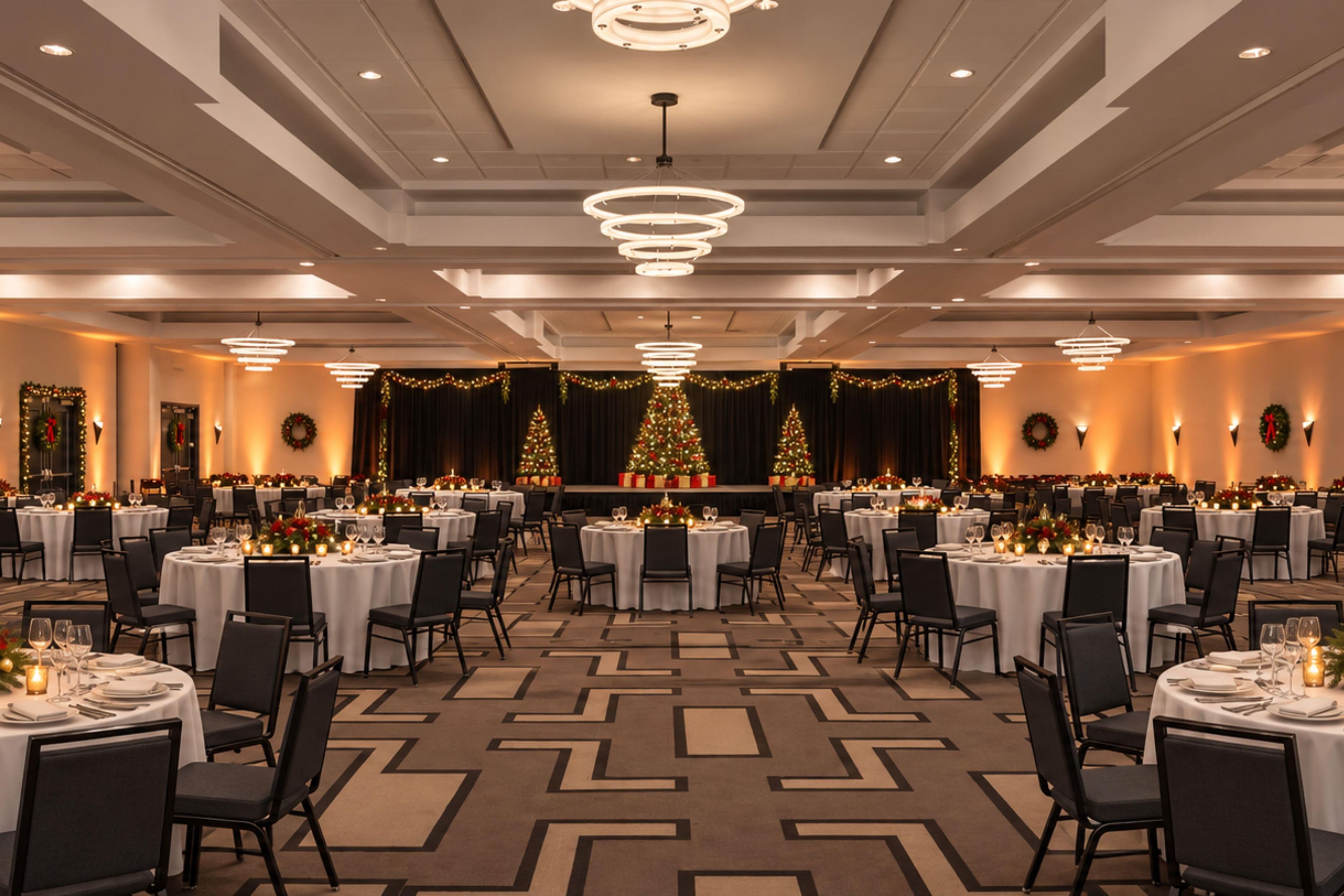 A banquet hall with round tables, chairs, and a central stage with Christmas trees.