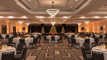A banquet hall with round tables, chairs, and a central stage with Christmas trees.