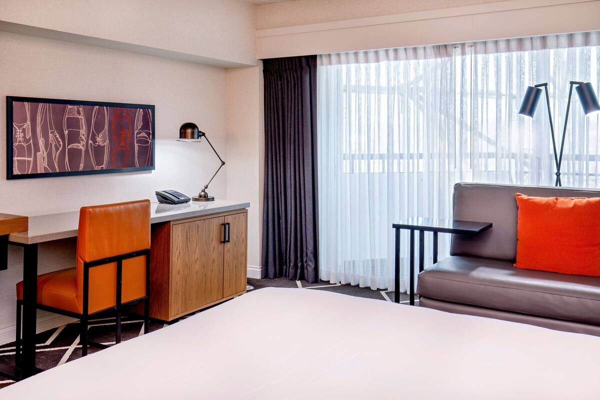 A hotel room with a bed, a desk with a chair, a sofa with an orange pillow, a painting on the wall, and a window with curtains.