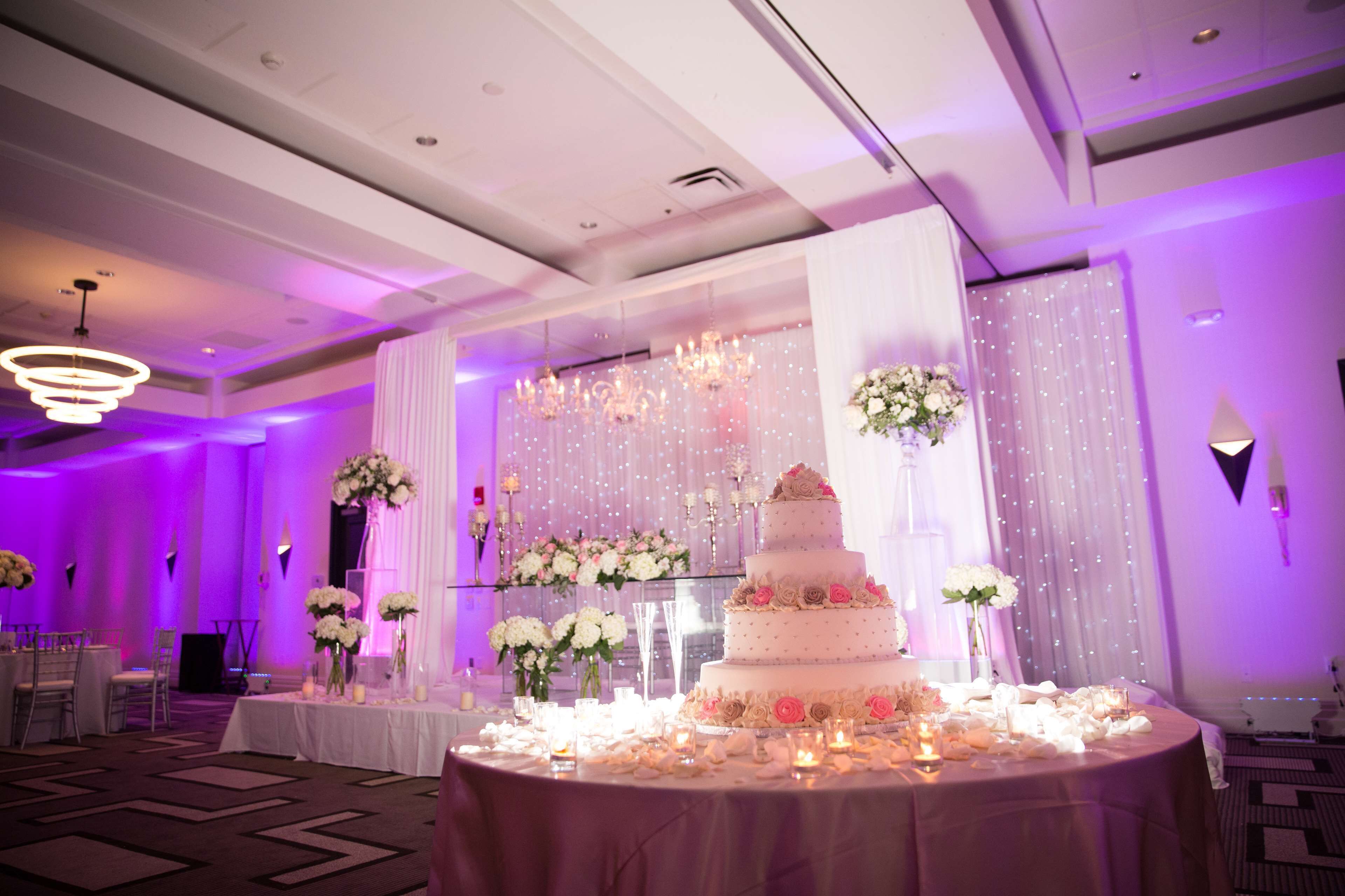 Salón de recepción de bodas con un pastel de varios niveles, centros de mesa florales e iluminación ambiental.