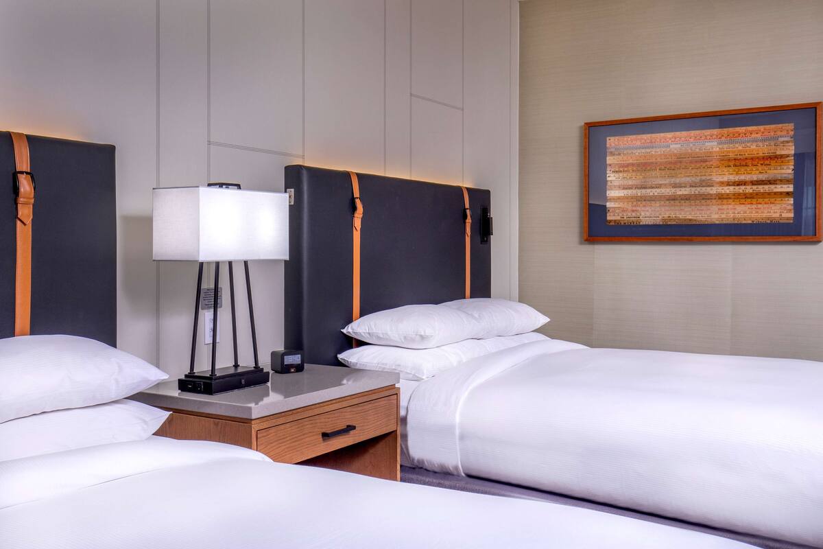 A hotel room with two beds, a bedside table with a lamp, and a framed artwork on the wall.
