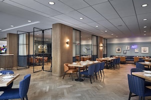 A modern restaurant interior with wooden flooring, tables with blue and brown chairs, and a glass partition wall.