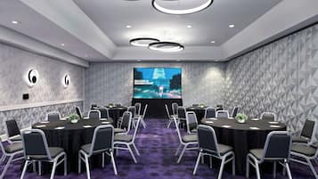 Un salón de conferencias con una pantalla grande que muestra “BIENVENIDOS A WASHINGTON DC”, una mesa redonda con sillas y un suelo cubierto de alfombra morada.
