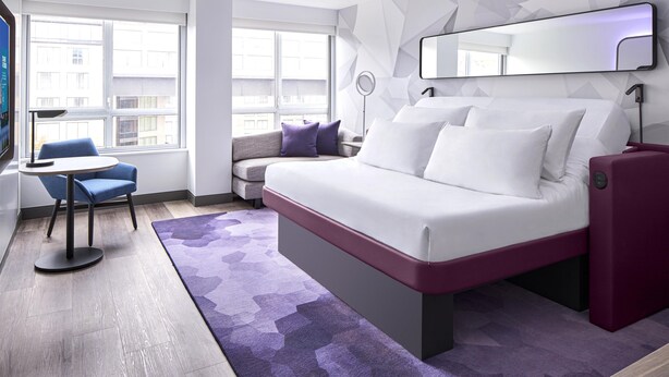 A modern hotel room with a bed, a desk, a chair, and a large window.
