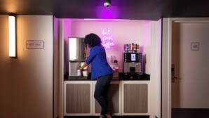 A woman in a blue shirt and jeans using a coffee machine in a modern kitchen area with a neon sign that reads "but first, coffee." - YOTEL Washington DC (Washington)