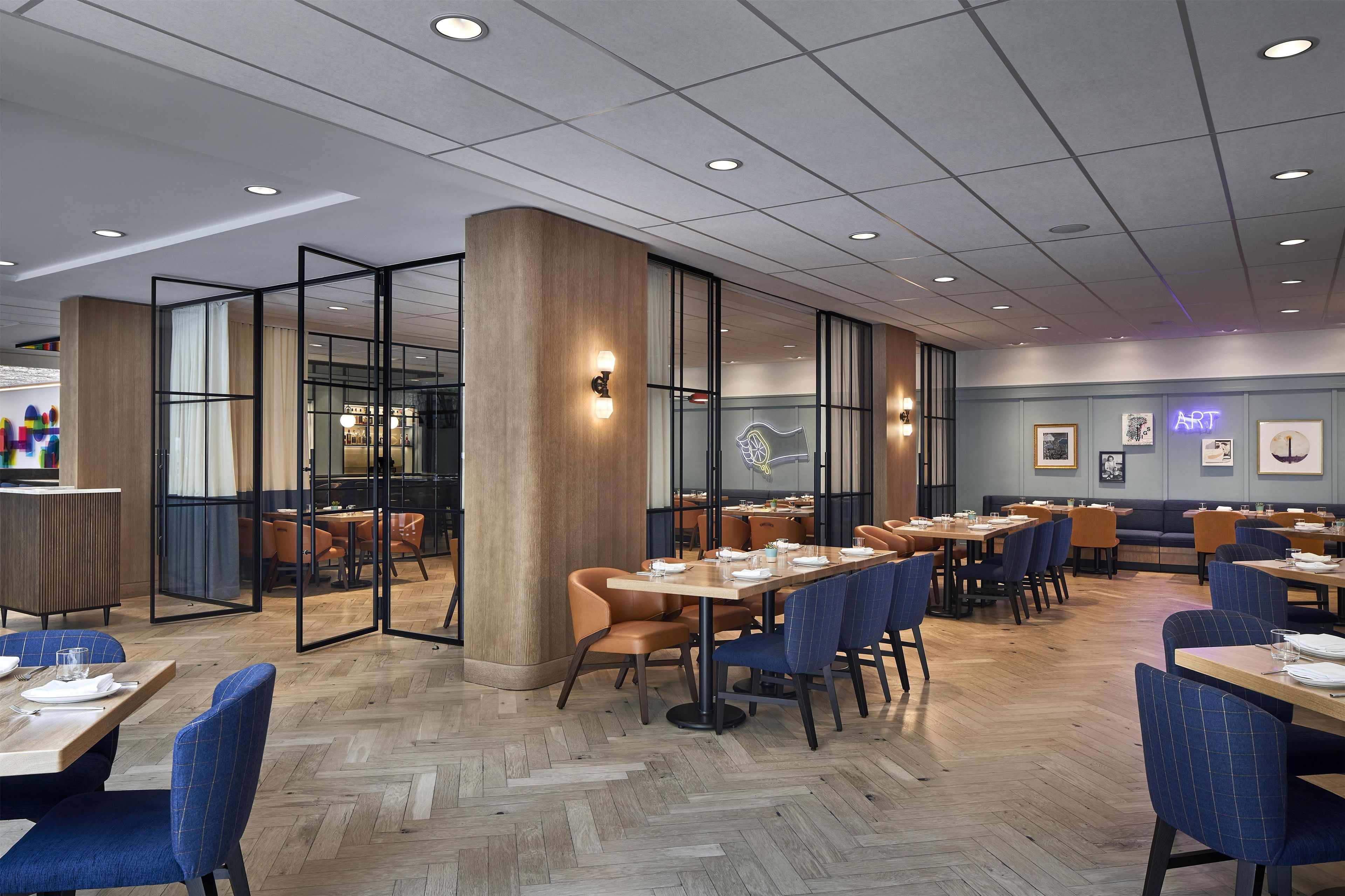 A modern restaurant interior with wooden flooring, tables with blue and brown chairs, and a glass partition wall.