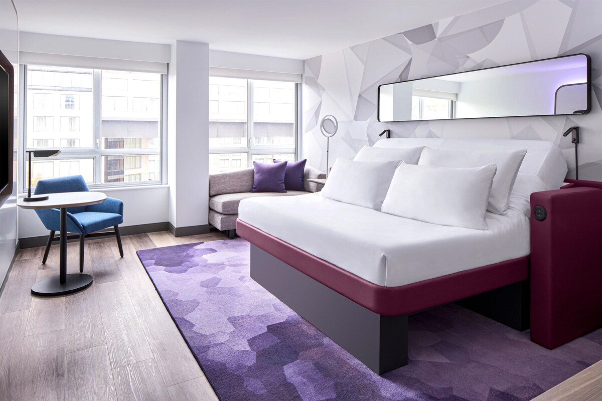 A modern hotel room with a bed, a desk, a chair, and a view of the city through large windows.
