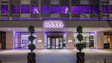 Entrada de hotel con un letrero de neón, puertas de vidrio y plantas en macetas.