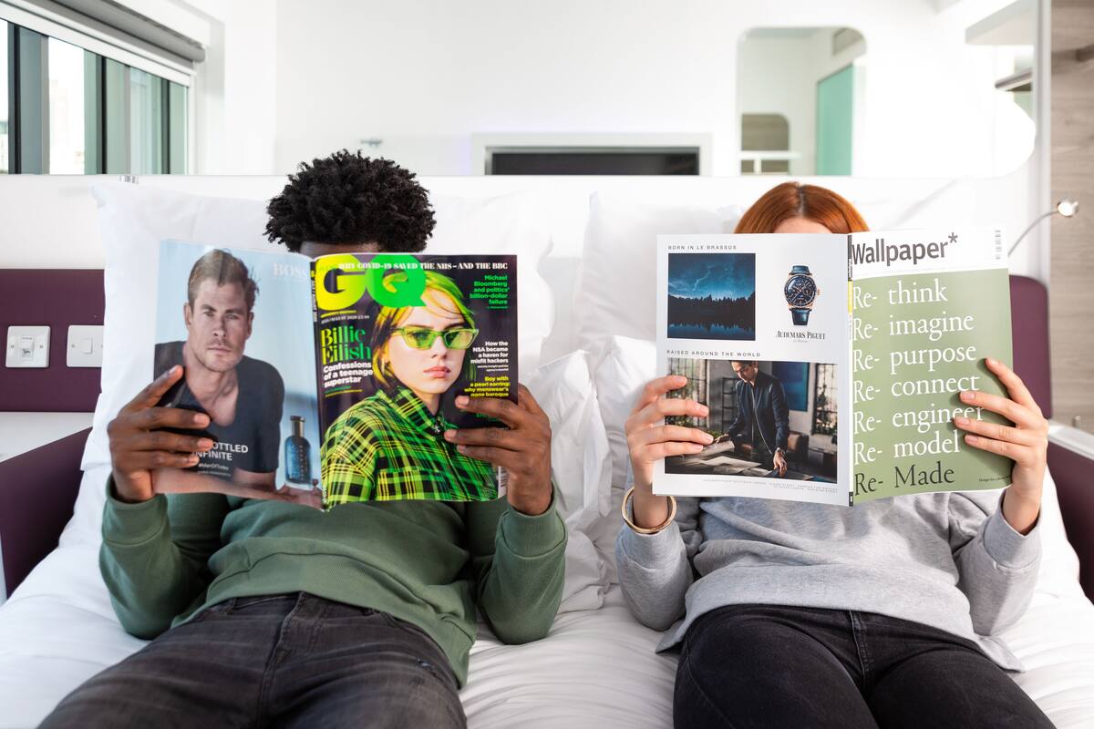 Dos personas recostadas en una cama, cada una sosteniendo una revista. La habitación tiene un diseño moderno con paredes blancas y una pared de acento púrpura.