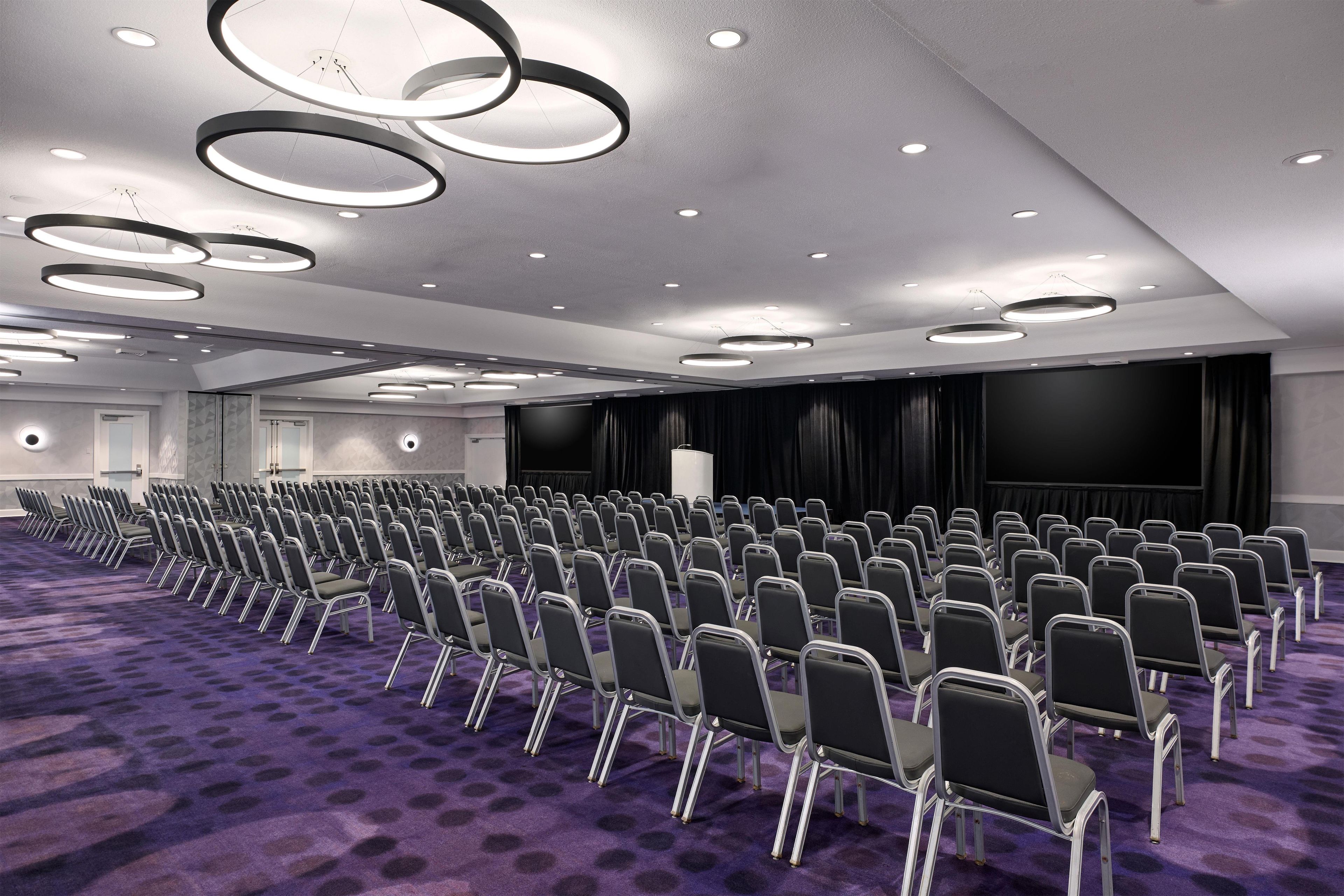A conference room with rows of chairs, a stage with curtains, and circular ceiling lights.