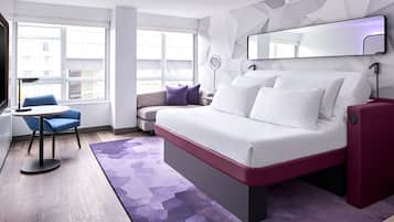 Een moderne hotelkamer met een bed, een bureau, een stoel en uitzicht op de stad door grote ramen.