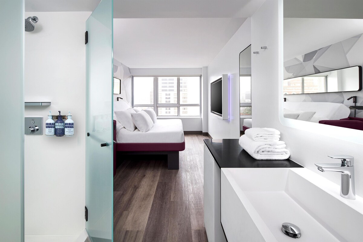 A modern hotel room with a bed, a large mirror, a bathtub, and a window with a city view.
