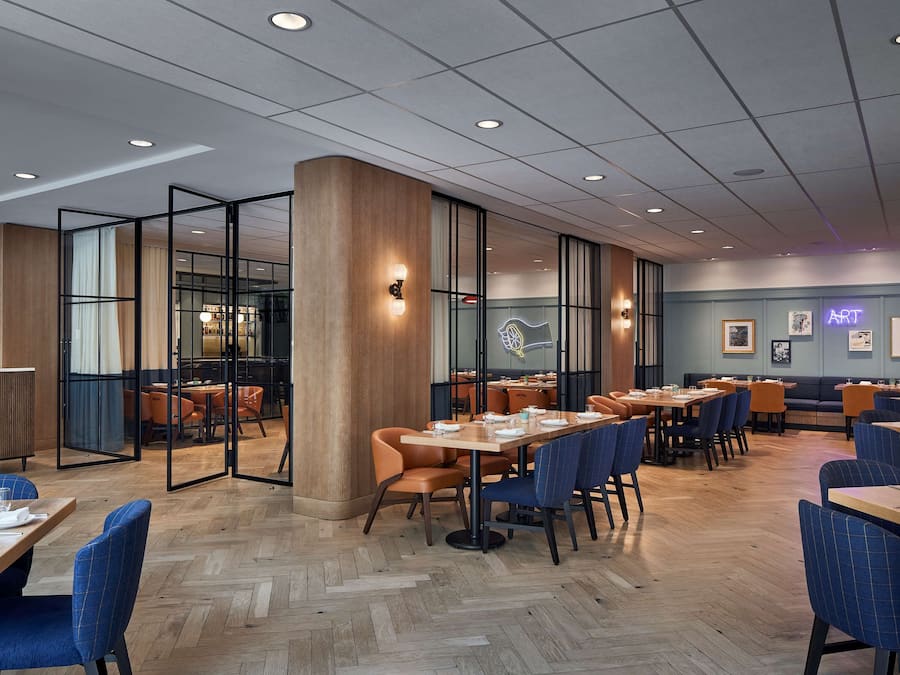 Ein modernes Restaurant-Interieur mit Holzboden, Glastrennwänden und unterschiedlichen Sitzmöglichkeiten, darunter Tische und Stühle.