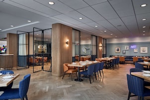 Interior de un restaurante moderno con suelo de madera, divisiones de vidrio y diferentes opciones de asientos, incluyendo mesas y sillas.