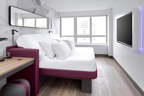 A modern hotel room with a bed, a desk, a chair, a TV, and a window with a city view. - YOTEL Washington DC, Select by Hilton (Washington)