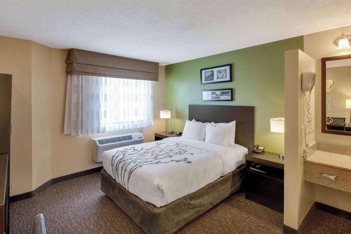 Une chambre d’hôtel avec un lit, une table de chevet, une lampe, un miroir et une fenêtre avec des rideaux.