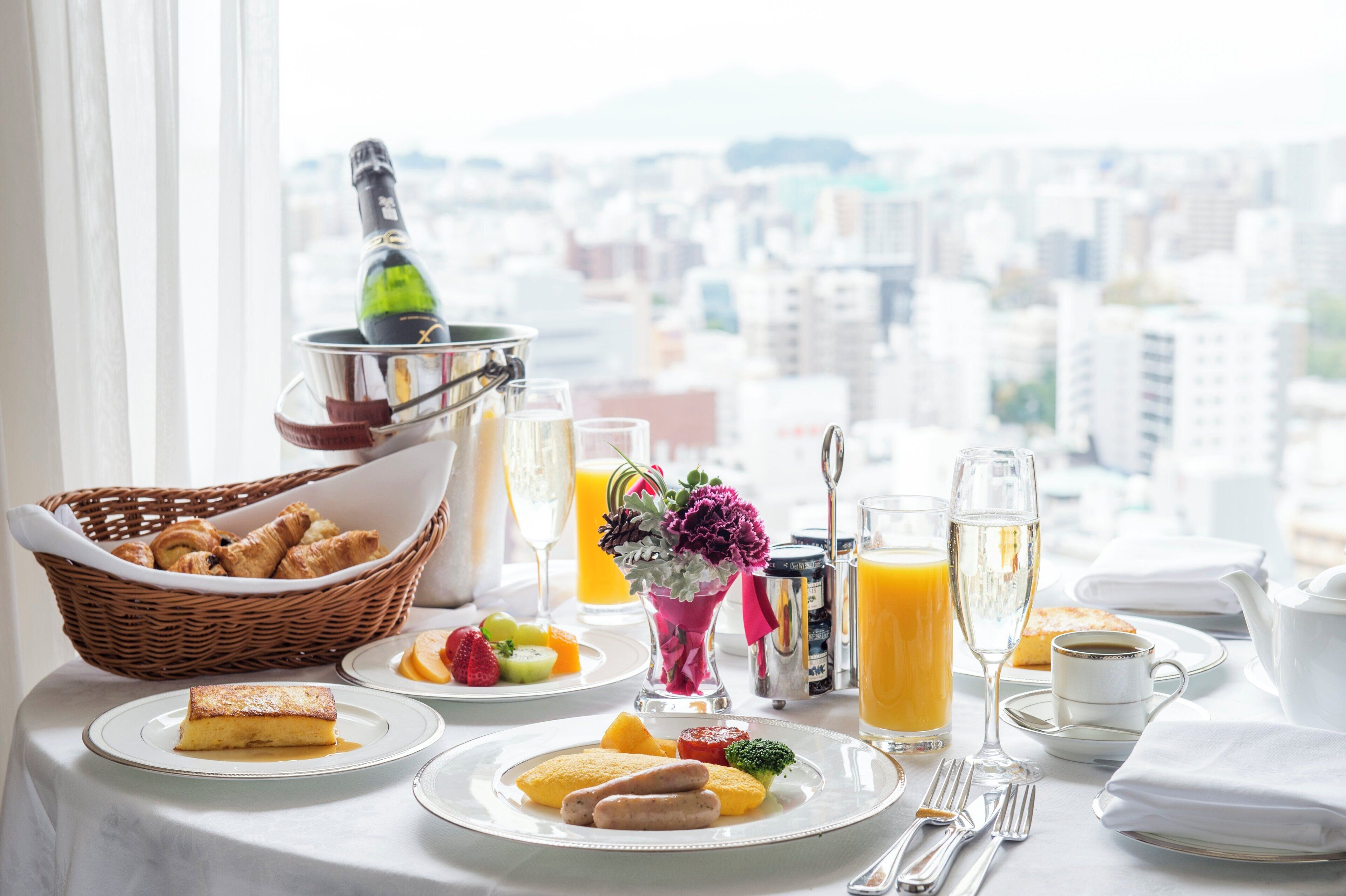 A breakfast table with croissants, fruit, and a bottle of champagne, set against a city view.