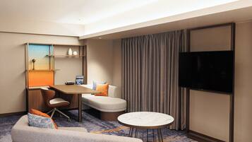 Habitación de hotel con sofá, una mesita, escritorio con silla, televisor y cortina.