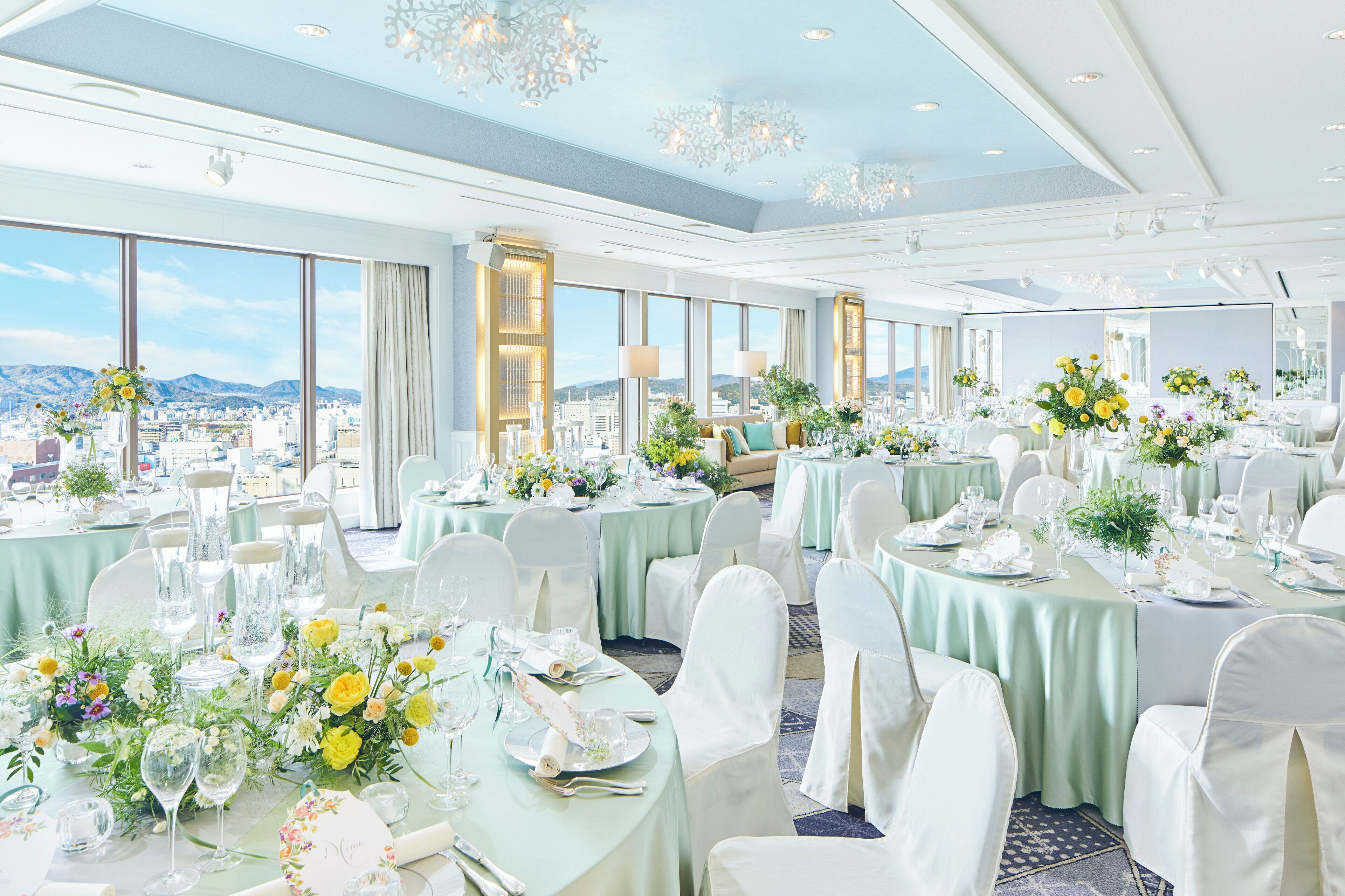A banquet hall with round tables set for a formal event, large windows offering a city view, and a high ceiling with chandeliers.