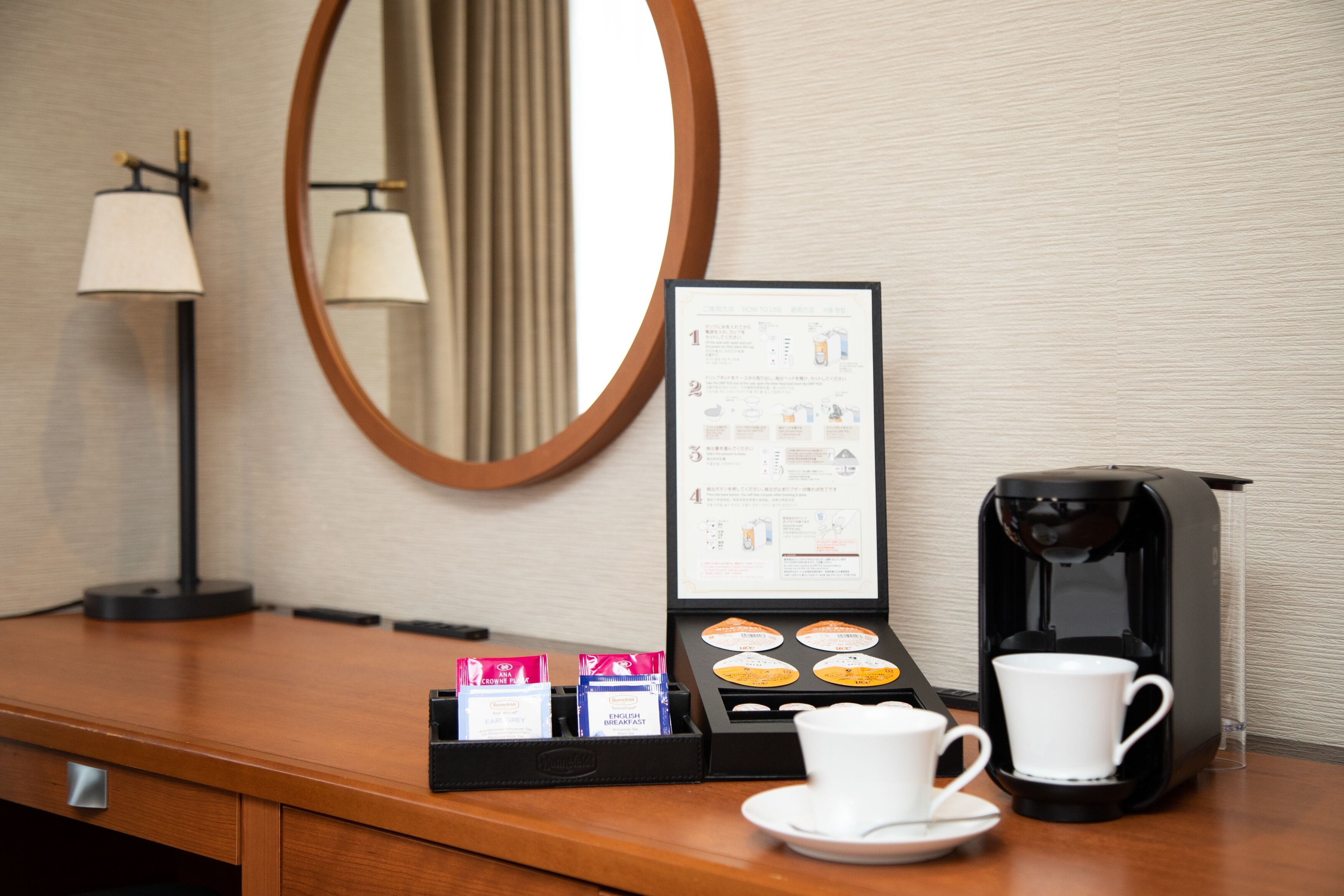 A hotel room with a wooden desk, a coffee maker, a cup, a tray with coffee pods, and a menu stand.