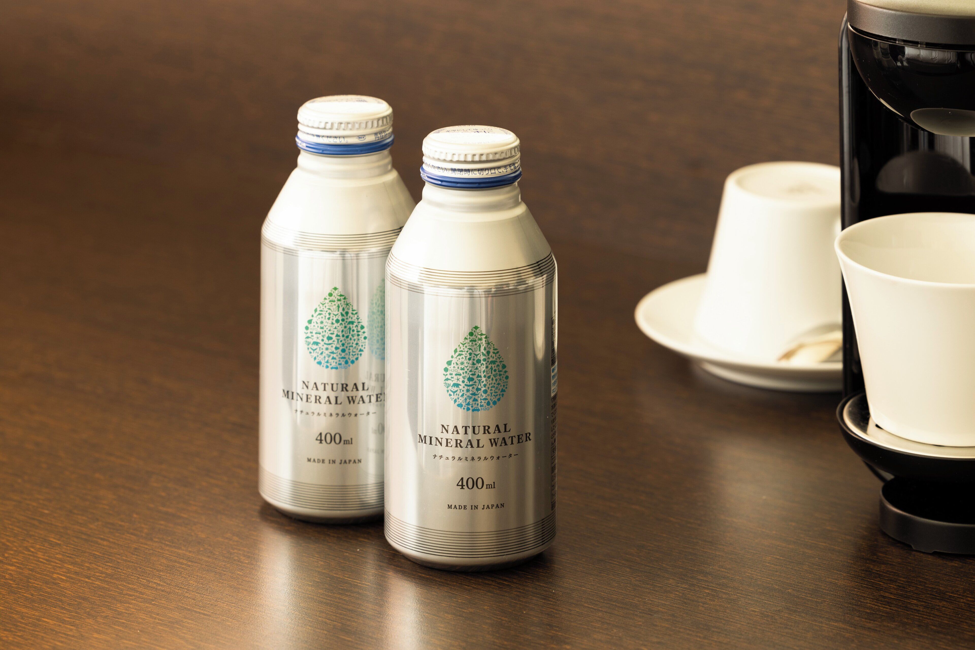 Dos botellas de agua mineral natural sobre una mesa de madera, junto con tazas y platillos blancos.