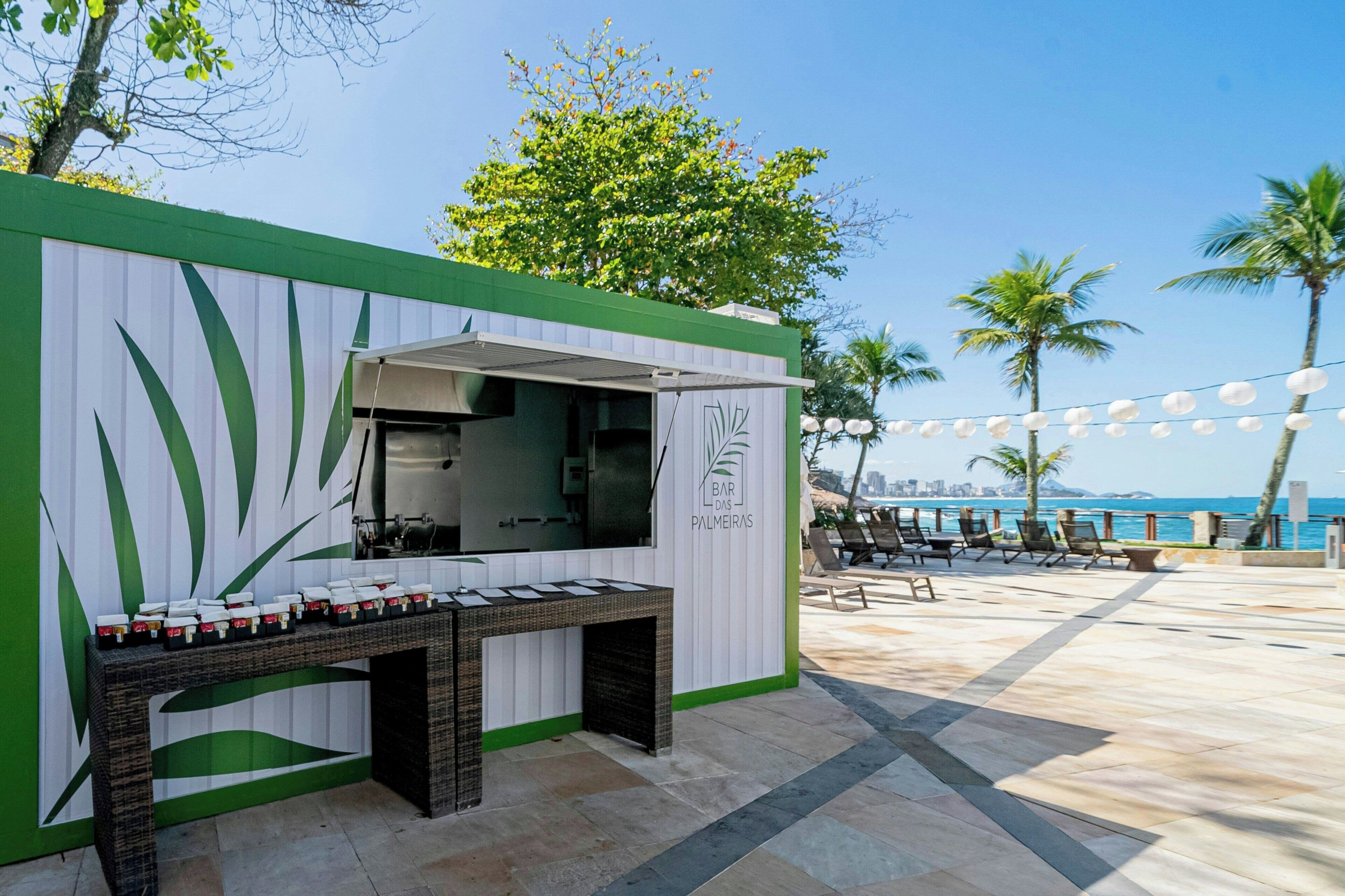 A small outdoor bar area with a counter and seating, surrounded by palm trees and a view of the city skyline.
