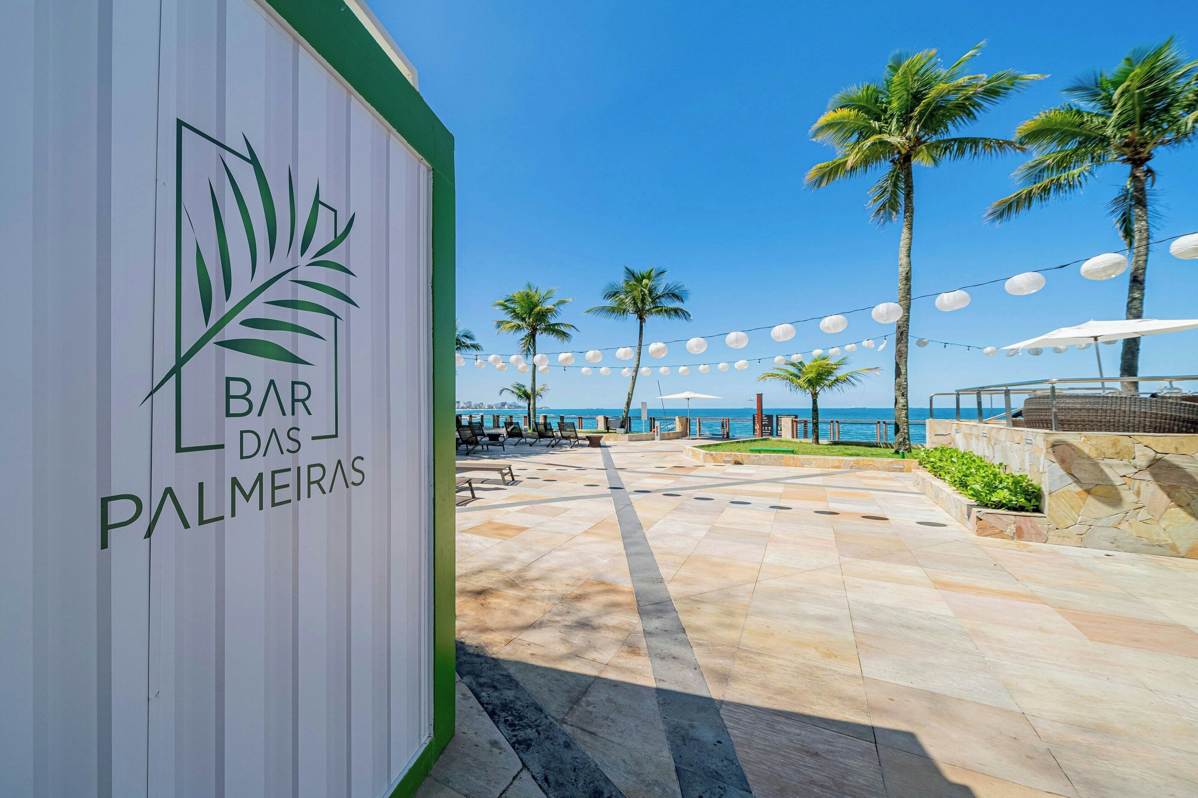 Un patio frente a la playa con un letrero que dice 'Bar das Palmeiras', palmeras y luces colgantes.