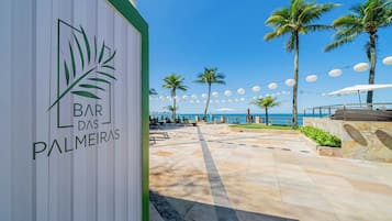 Un patio frente a la playa con un letrero que dice 'Bar das Palmeiras', palmeras y luces colgantes.