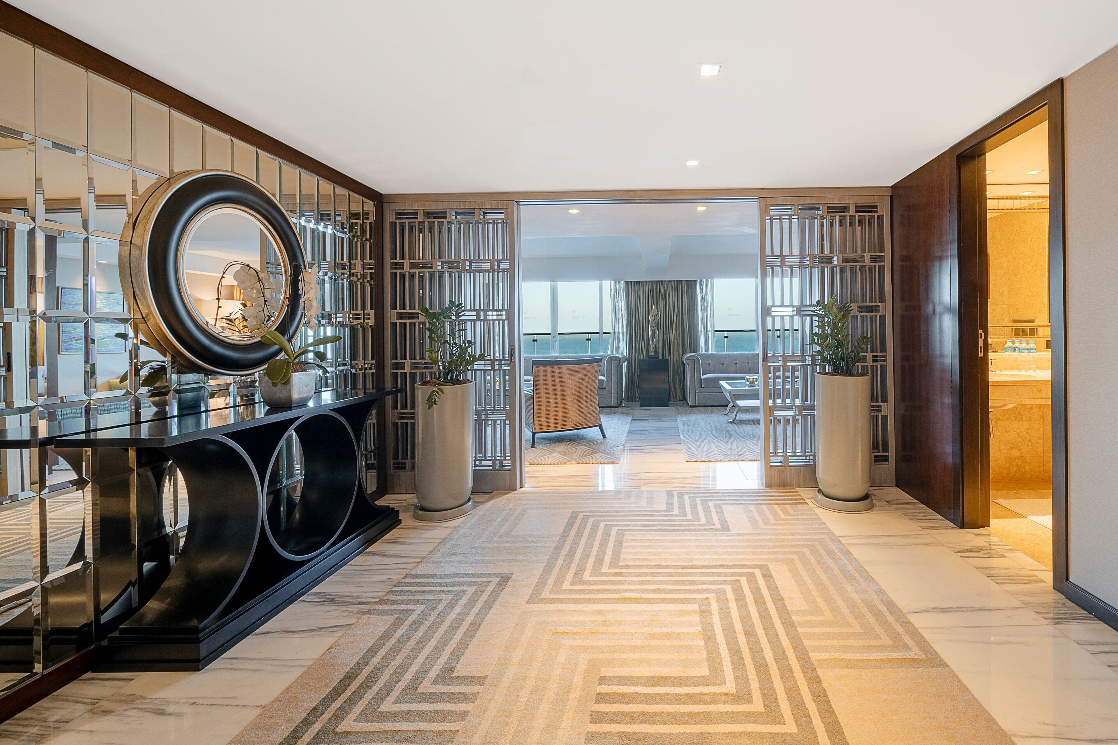 Un hall d’hôtel moderne avec un sol en marbre, une console noire surmontée d’un grand miroir et une vue sur l’espace de vie à travers une porte en verre.